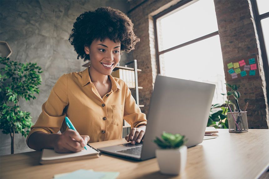 Photo of smiling African American woman using a laptop and writing on a notepad